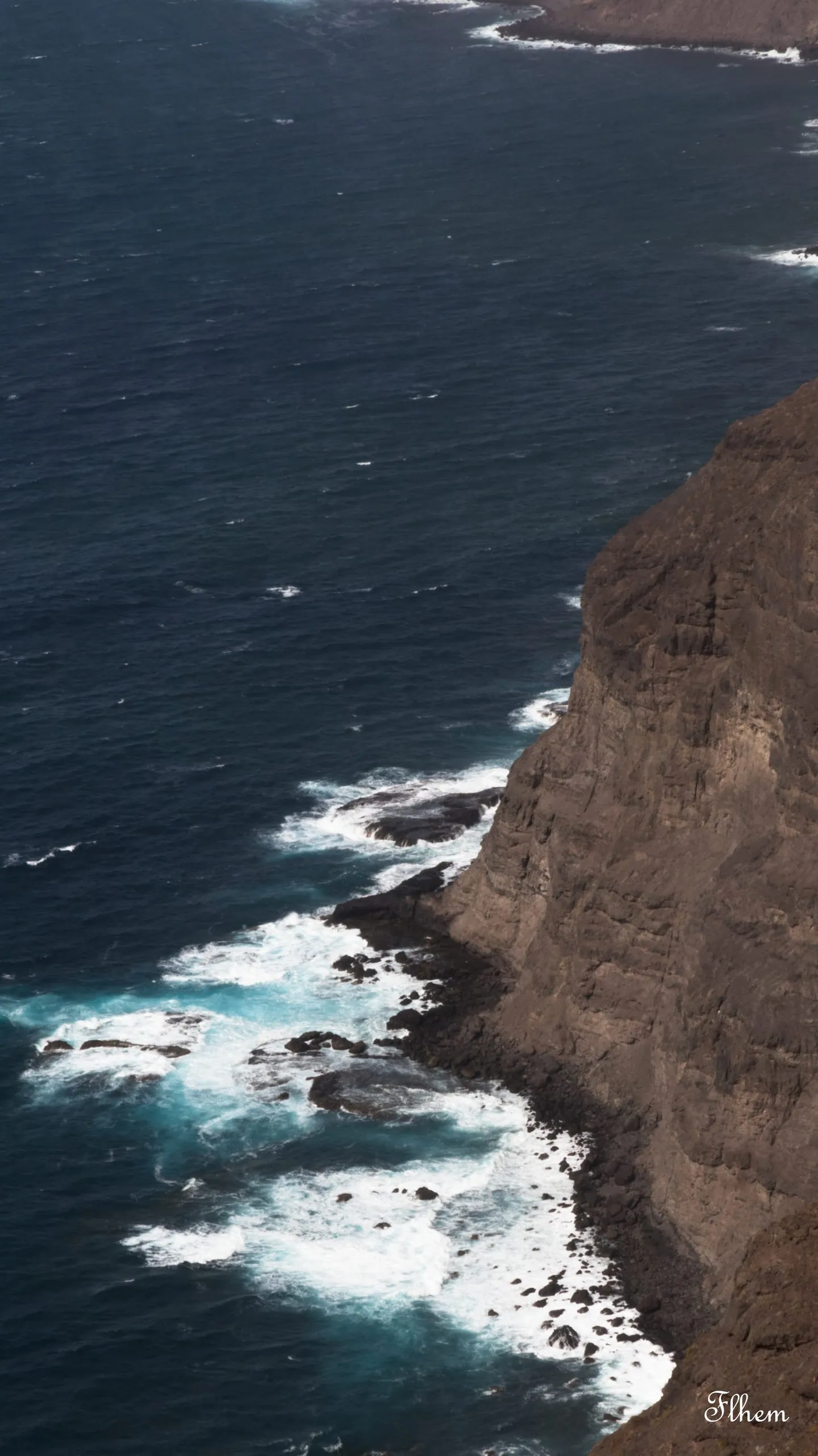 Falaise abrupte plongeant dans une mer bleu sombre frangée d'écume, vue d'en haut