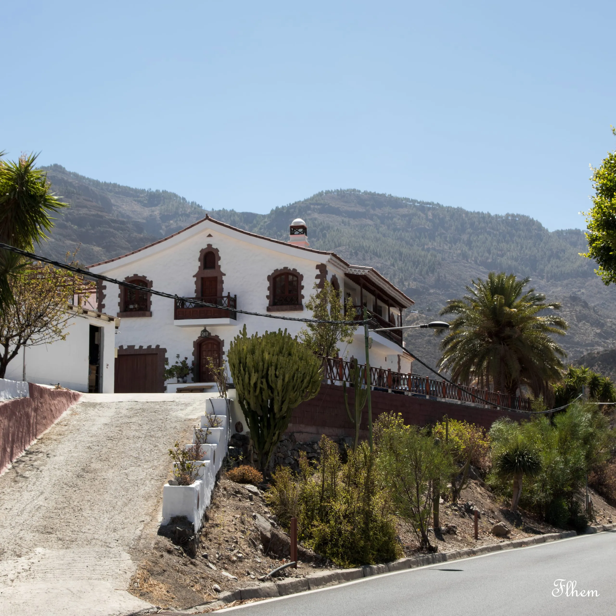 Maison blanche aux encadrements bruns en haut d'une allée en pente, cactus et palmiers, montagnes derrière