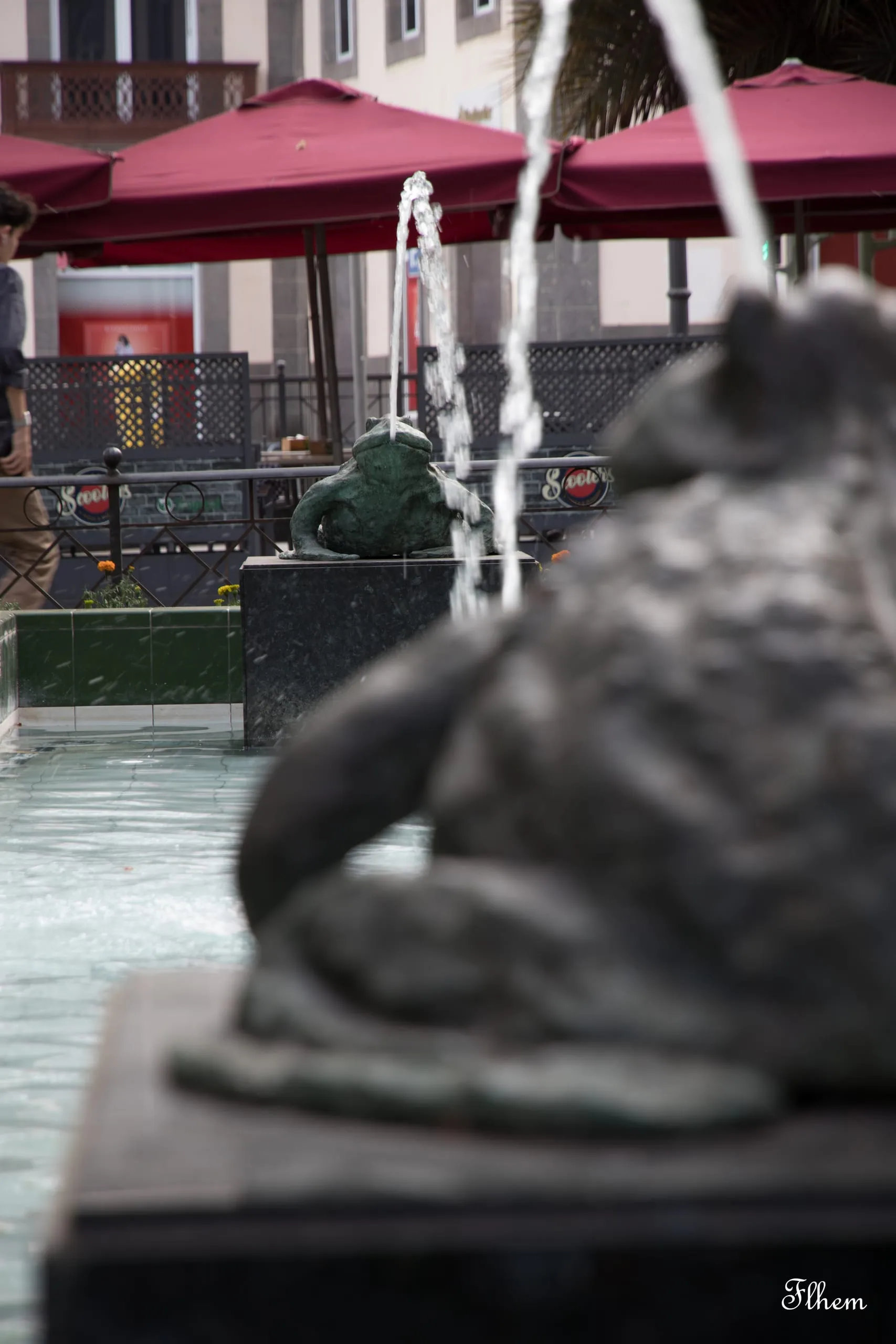 Statue de grenouille dans une fontaine, parasols rouges d'une terrasse en arrière-plan